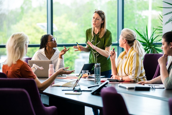 Group of women in a business meeting at the workplace, with the leader standing up and presenting an idea to the rest. 