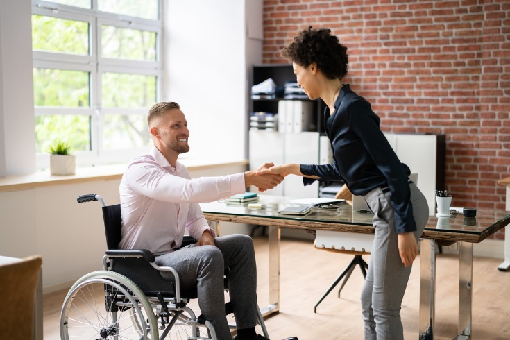 Business woman shaking disabled or injured colleague’s hand who is in a wheelchair in the office.