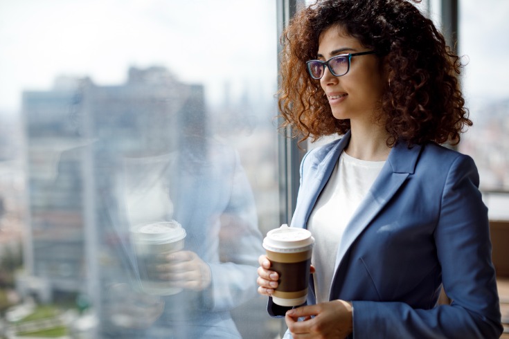 Businesswoman looks out the window of her office while holding a coffee and practicing corporate mindfulness.