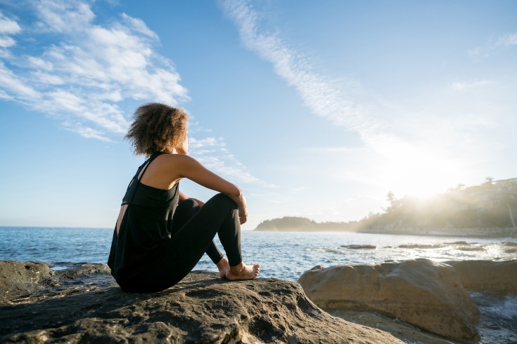 Woman sitting on the rocks at a beach while practicing mindfulness and meditation.