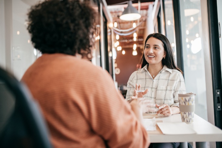 A woman speaking with another woman across a table in a casual workplace setting with coffee and paperwork in front of them.