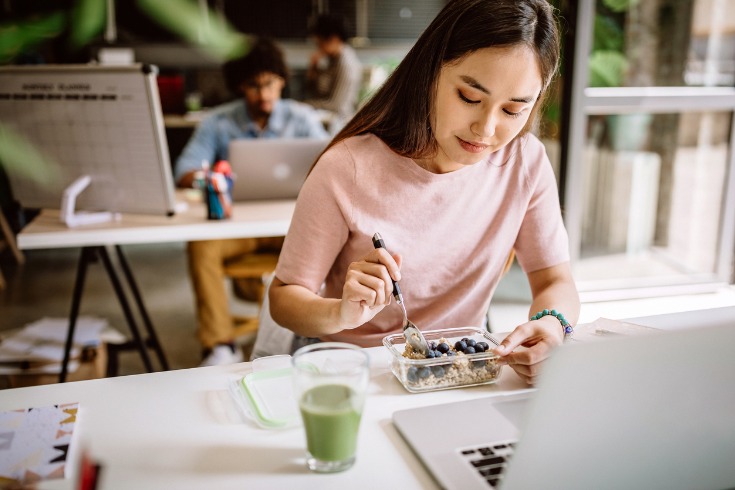 Woman eating a nutritious breakfast while sitting at her desk to help with staying healthy at work.